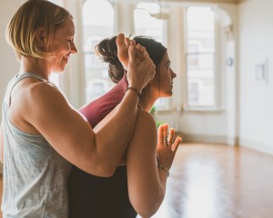 Ashtanga yoga teacher Arielle Nash helping Siobhan Sears put leg behind head while standing